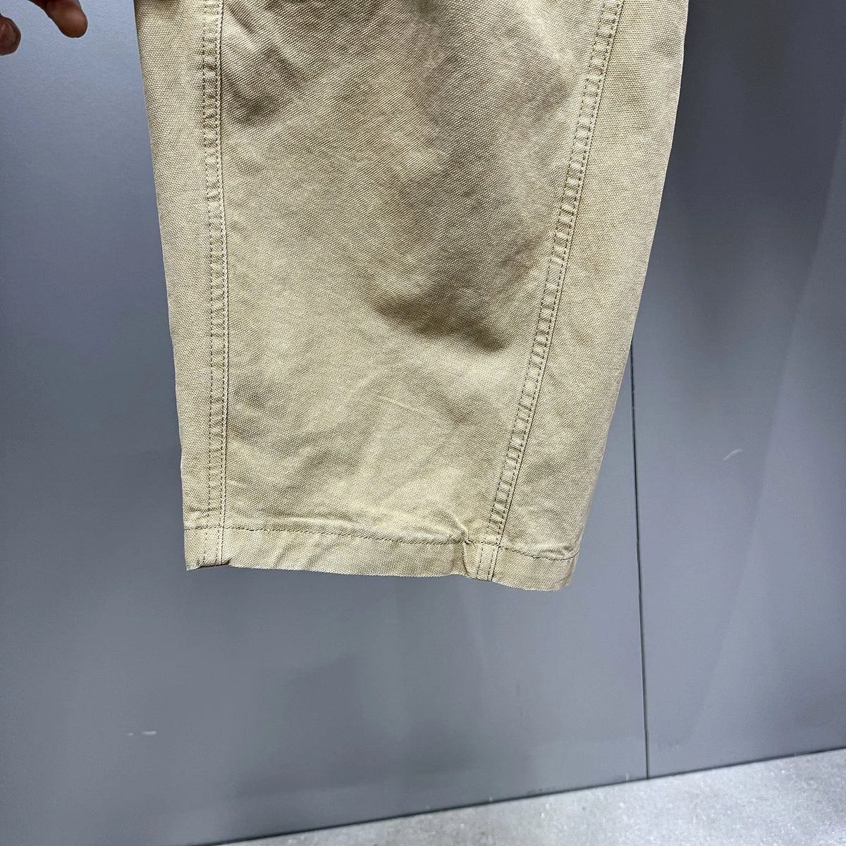 A close-up of a light beige or khaki fabric garment, showing the texture and hem stitching, held against a gray background.