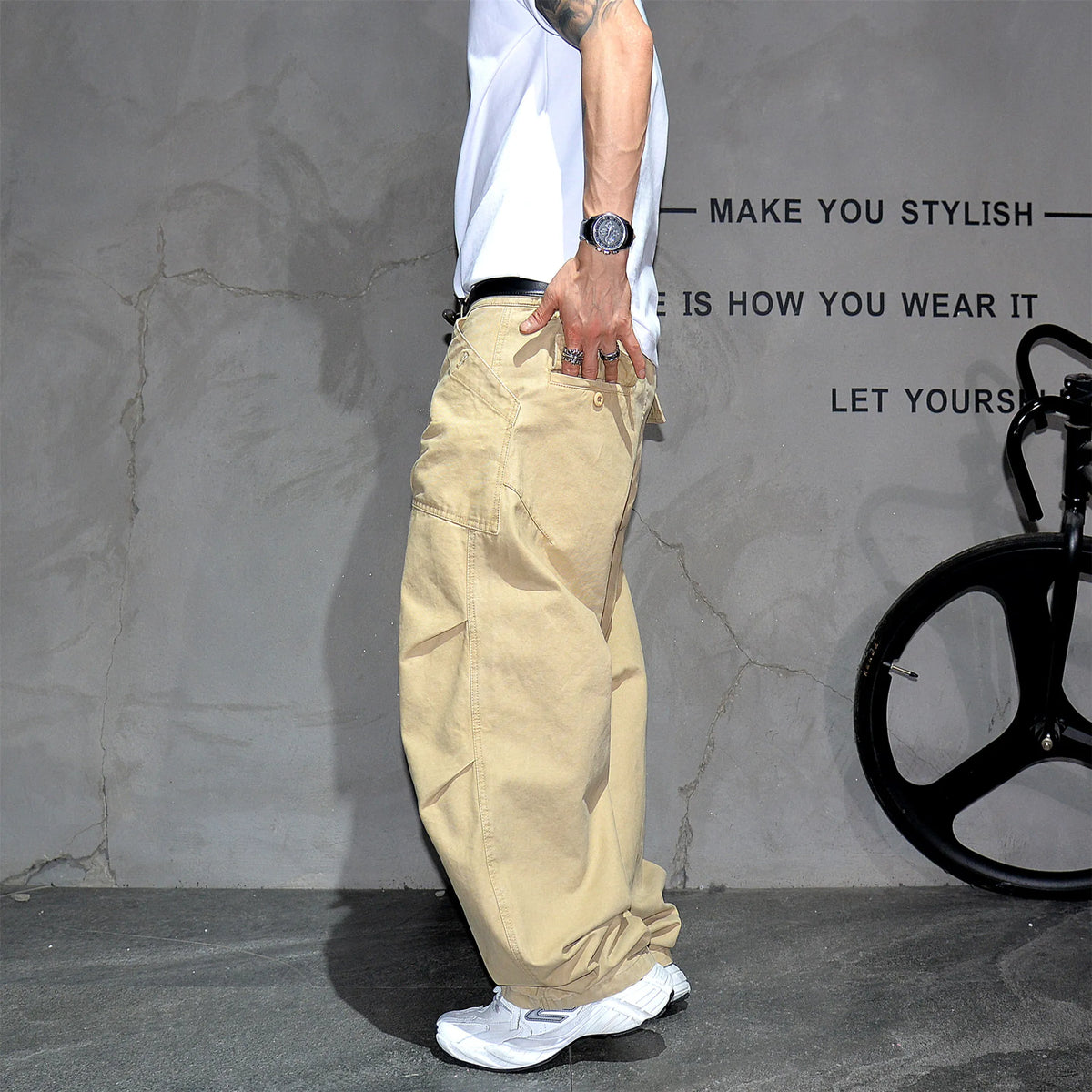 A person wearing loose beige cargo pants, a white t-shirt, and white sneakers stands in front of a textured gray wall with black text and a bicycle wheel visible on the right.