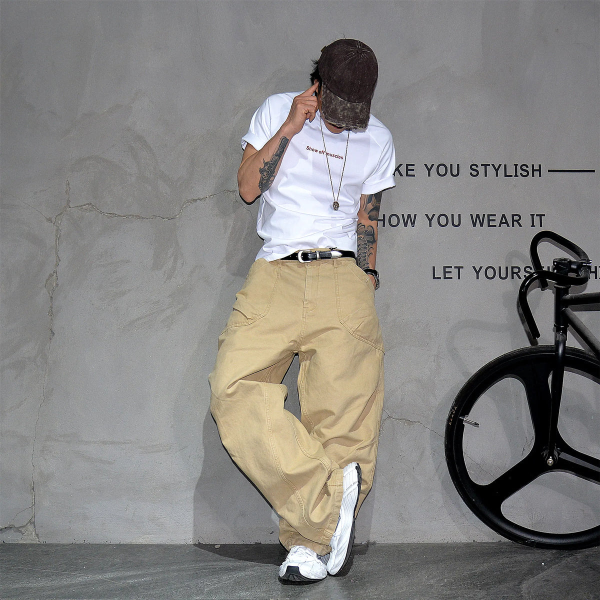 A person wearing a white graphic T-shirt, beige wide-leg cargo pants, a dark cap, and white sneakers poses against a textured gray wall with a black bicycle wheel visible on the right.
