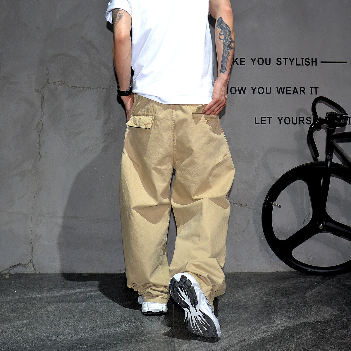 A person from the back wearing baggy beige cargo pants, a white t-shirt, and black-and-white sneakers, standing against a textured gray wall with a black bicycle wheel visible on the right.