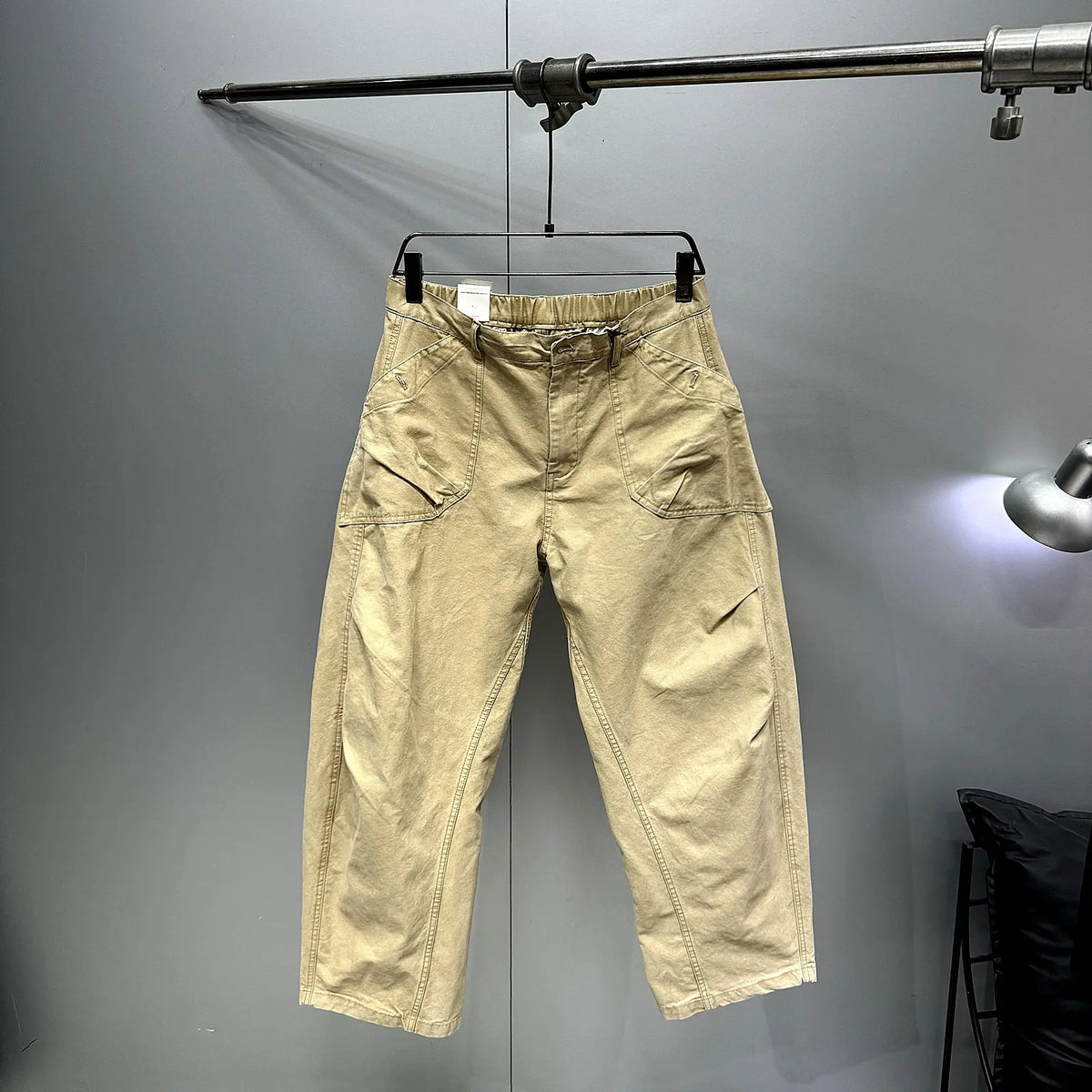 A pair of beige cargo-style pants hanging on a black hanger from a metal clothing rack against a gray wall.